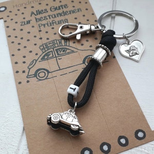 Könnte beinhalten: Schwarzer und silberner Schlüsselanhänger mit einem kleinen silbernen Auto-Anhänger, einem silbernen Engel-Anhänger und einer schwarz-weißen Perle mit dem Buchstaben "T". Der Schlüsselanhänger ist an einer Karte mit dem Text "Alles Gute zur bestandenen Prüfung" befestigt.