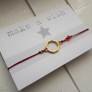 Könnte beinhalten: Rotes Armband mit einem goldenen sechseckigen Anhänger und einer roten Kristallperle. Das Armband liegt auf einer weißen Karte mit dem Text "make a wish" und einem silbernen Stern.