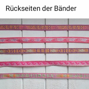 May include: Five different woven ribbon trims with various patterns and colours, including pink, purple, orange, and gold. The ribbons are displayed on a white wooden surface. The text "R&uuml;ckseiten der B&auml;nder" is visible at the top of the image.