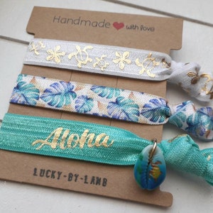 Könnte beinhalten: Set aus drei Haargummis mit tropischem Thema. Die Haargummis sind weiß mit goldenen Blumendesigns, weiß mit blauen und grünen tropischen Blattdesigns und türkis mit goldener "Aloha"-Beschriftung. Das türkisfarbene Haargummi hat einen kleinen Muschel-Anhänger.