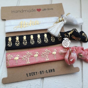 Könnte beinhalten: Set aus drei elastischen Haargummis mit goldenen Akzenten. Die Haargummis haben ein weißes Band mit dem Wort "Aloha" in goldenen Buchstaben, ein schwarzes Band mit goldenen Ananas-Designs und ein rosa Band mit goldenen Palmen und Flamingos. Die Haargummis sind auf einer braunen Karte mit dem Text "Handmade with love" und "Lucky-By-Lamb" verpackt.