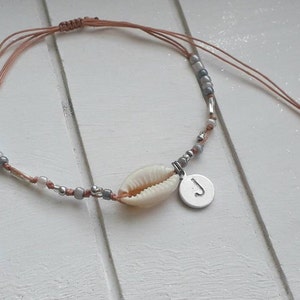 Könnte beinhalten: Ein zartes Armband mit einem Muschel-Anhänger und einem silbernen Anhänger mit dem Buchstaben 'J'. Das Armband ist aus braunen und grauen Perlen und einer braunen Schnur gefertigt.