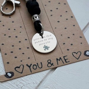 Könnte beinhalten: Ein silberner Schlüsselanhänger mit einem schwarzen Band und einem silbernen Anhänger, auf dem "You are the piece to my puzzle" mit einem Puzzle-Symbol steht. Der Schlüsselanhänger befindet sich auf einer braunen Karte mit den Worten "You & Me", die mit einem schwarzen Stift geschrieben wurden.