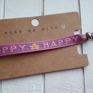 May include: A purple and pink woven bracelet with the words "Happy" in pink and a yellow flower design. The bracelet has a silver metal clasp and a brown card tag that says "Wear me with &hearts;".