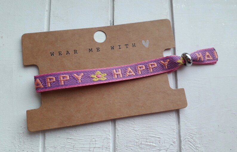 May include: A purple and pink woven bracelet with the words "Happy" in pink and a yellow flower design. The bracelet has a silver metal clasp and a brown card tag that says "Wear me with &hearts;".