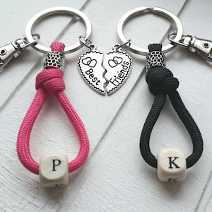 Könnte beinhalten: Zwei beste Freundinnen Schlüsselanhänger-Sets mit silbernen Herzanhängern, auf denen "Best" und "Friends" steht. Ein Schlüsselanhänger ist pink mit einer Perle mit dem Buchstaben "P" und der andere ist schwarz mit einer Perle mit dem Buchstaben "K".