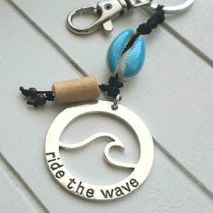 Könnte beinhalten: Silberner Schlüsselanhänger mit einem Wellendesign und dem Text "Ride the Wave". Der Schlüsselanhänger enthält auch eine blaue Muschel und braune Holzperlen.