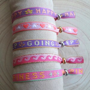 May include: Five colourful woven friendship bracelets with gold and silver accents. The bracelets feature the words "Happy", "Keep Going", "Happiness", and "Happy" with hearts and stars.