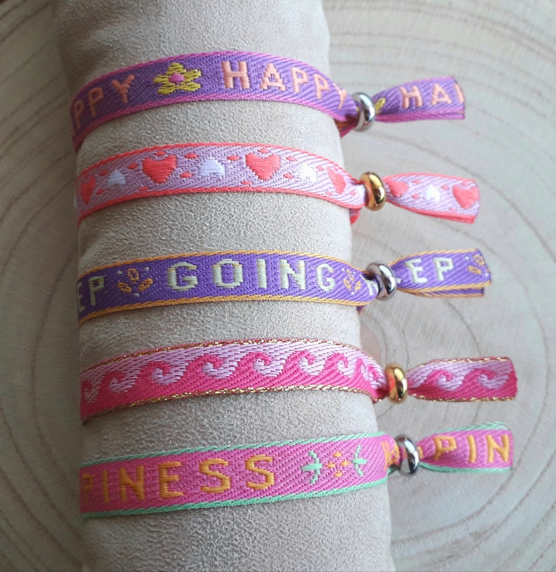 May include: Five colourful woven friendship bracelets with gold and silver accents. The bracelets feature the words "Happy", "Keep Going", "Happiness", and "Happy" with hearts and stars.