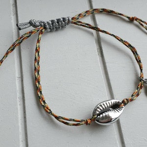 Könnte beinhalten: Ein mehrfarbiges geflochtenes Armband mit einem silbernen Muschel-Anhänger und einem silbernen Palmen-Anhänger.
