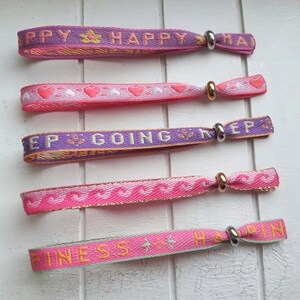 May include: Five colourful woven wristbands with different messages, including "Happy", "Keep Going", and "Happiness". Each band has a silver metal clasp.