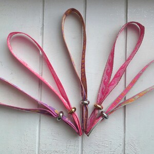 May include: Five colourful fabric wristbands with silver metal rings. Each wristband has a different pattern and colour combination, including pink, purple, and orange.