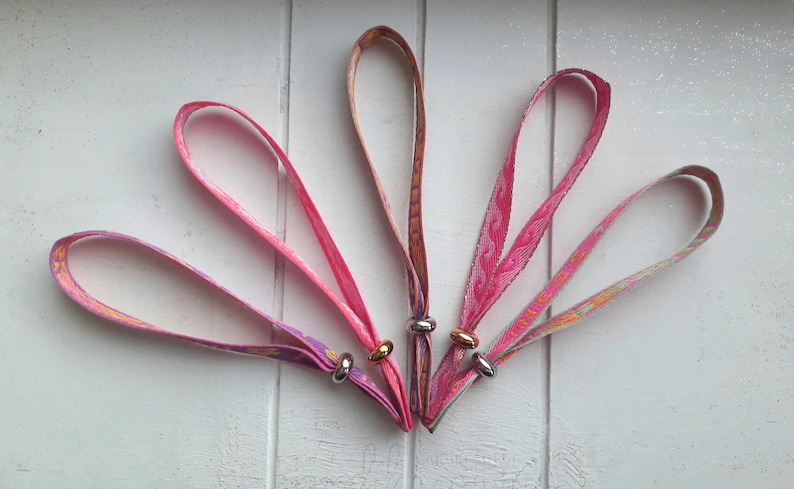 May include: Five colourful fabric wristbands with silver metal rings. Each wristband has a different pattern and colour combination, including pink, purple, and orange.