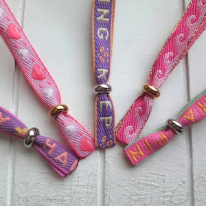 May include: Five colourful fabric wristbands with silver and gold metal clasps. The wristbands have the words "Happy", "Ninja", "Sing", "Keep" and "With" printed on them.