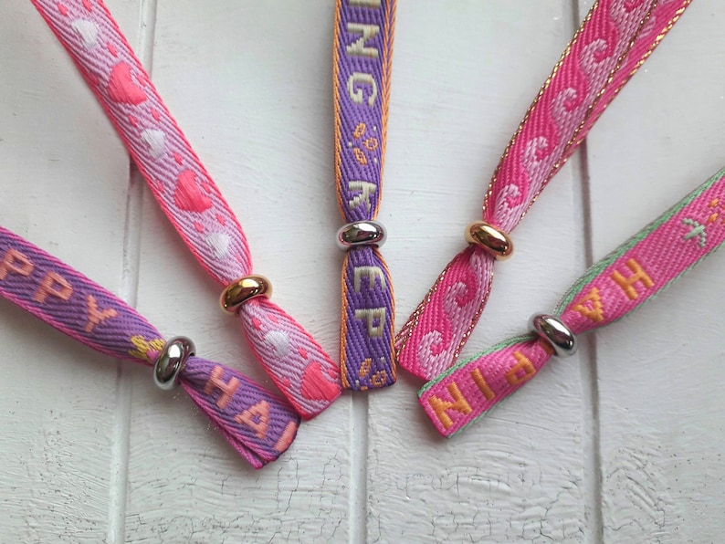 May include: Five colourful fabric wristbands with silver and gold metal clasps. The wristbands have the words "Happy", "Ninja", "Sing", "Keep" and "With" printed on them.