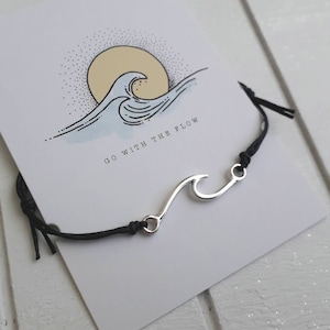 Könnte beinhalten: Schwarzes Armband aus Schnur mit einem silbernen Wellen-Anhänger. Das Armband hat an einem Ende einen Knoten und am anderen einen Haken. Der Anhänger ist eine stilisierte Welle mit einem Haken am Ende. Das Armband liegt auf einer weißen Karte mit dem Text "Go with the flow" und einer Zeichnung einer Welle und der Sonne.