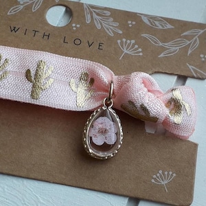 Könnte beinhalten: Rosa elastisches Haargummi mit goldfarbenem Kaktusdruck und einem goldenen Anhänger mit einer gepressten Blume darin. Die Verpackung trägt den Schriftzug "With Love".