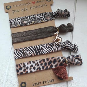 Könnte beinhalten: Satz mit fünf Haargummis mit Tiermuster. Die Haargummis haben ein Zebra-Muster, ein Leopardenmuster und ein braunes Haargummi mit einem goldenen Zebra-Anhänger. Die Haargummis sind auf einer braunen Karte verpackt mit dem Text "Handmade with love You are amazing" und "Lucky-By-Lamb".