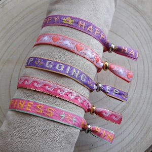 May include: Five colourful friendship bracelets with different messages. The bracelets are pink, purple, and green with gold and silver accents. The messages on the bracelets are "Happy", "Keep Going", "Happiness", and "Happy".