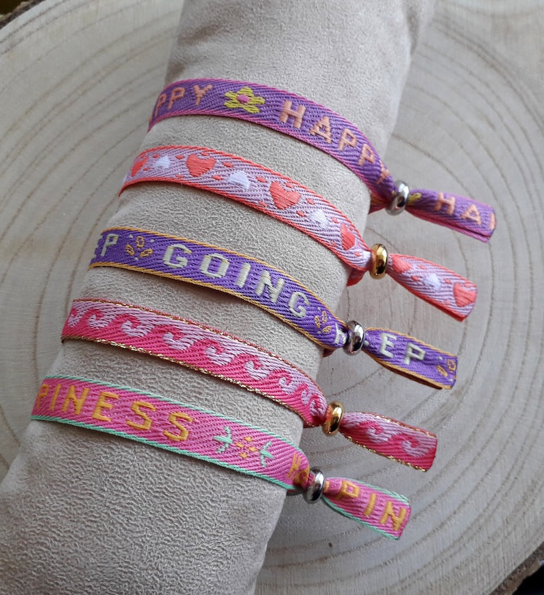 May include: Five colourful friendship bracelets with different messages. The bracelets are pink, purple, and green with gold and silver accents. The messages on the bracelets are "Happy", "Keep Going", "Happiness", and "Happy".