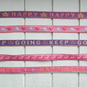 May include: Five different pink and purple ribbon trims with various patterns and text. The text on the ribbons reads "Happy", "Keep Going", and "Happiness".