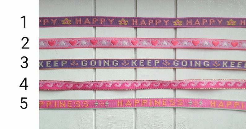 May include: Five different pink and purple ribbon trims with various patterns and text. The text on the ribbons reads "Happy", "Keep Going", and "Happiness".