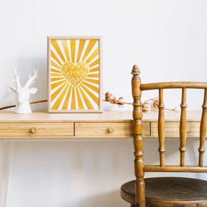 May include: A framed print with a yellow and white sunburst design and a heart with the text "You are my sunshine" in white. The print is on a wooden desk with a white deer figurine and a wooden chair.