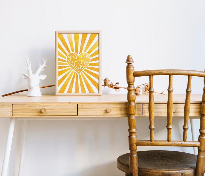 May include: A framed print with a yellow and white sunburst design and a heart with the text "You are my sunshine" in white. The print is on a wooden desk with a white deer figurine and a wooden chair.