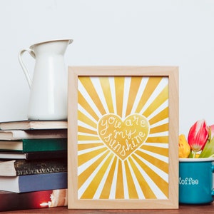 May include: A framed print with a yellow and white sunburst design. The print features a heart with the text "You are my sunshine" in white. The print is sitting on a table with a stack of books and a blue mug with the word "Coffee" on it.