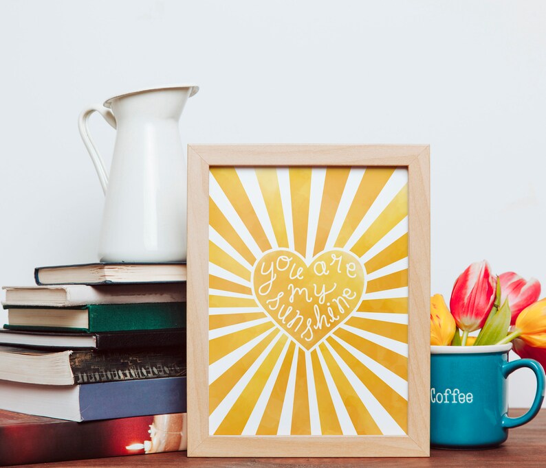 May include: A framed print with a yellow and white sunburst design. The print features a heart with the text "You are my sunshine" in white. The print is sitting on a table with a stack of books and a blue mug with the word "Coffee" on it.