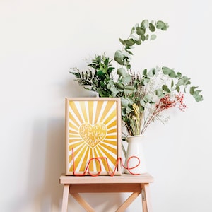 May include: A yellow and white framed print with a sunburst design and a heart that says "You are my sunshine". A red metal word "love" is in front of the print. A vase with green and brown flowers is on a wooden stool.