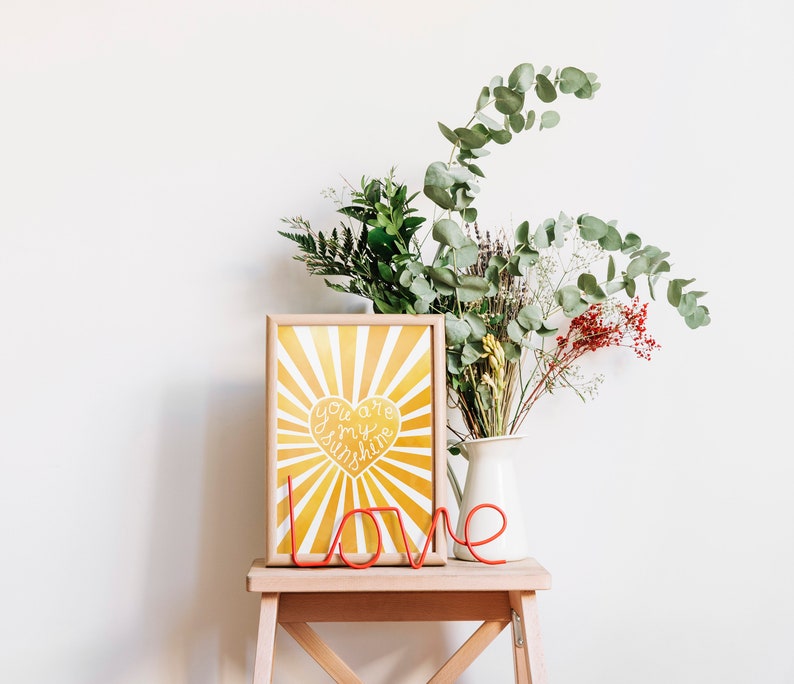 May include: A yellow and white framed print with a sunburst design and a heart that says "You are my sunshine". A red metal word "love" is in front of the print. A vase with green and brown flowers is on a wooden stool.