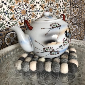 Può includere: Una teiera bianca con un motivo floreale e un manico rosso si trova su un sottobicchiere di feltro grigio e bianco. La teiera ha un motivo di uccello e fiori.