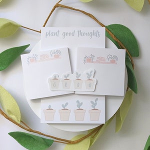 May include: A set of four white notepads with the text "plant good thoughts" on the top notepad. The notepads feature illustrations of potted plants and the word "GROW" in a sticker on the bottom notepad.