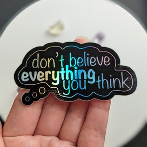 May include: A holographic sticker in the shape of a thought bubble. The sticker has a black border and contains the words "don't believe everything you think" in a rainbow gradient font. The sticker is held in a hand.