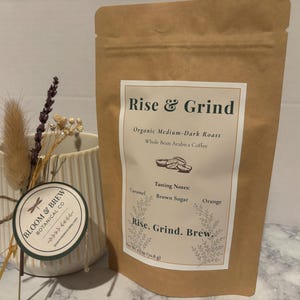 May include: A brown paper bag of "Rise & Grind" organic medium-dark roast whole bean Arabica coffee. The bag has a white label with tasting notes of caramel, brown sugar, and orange. A white mug with a floral arrangement is next to the bag.