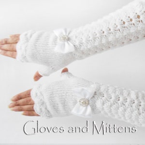 Könnte beinhalten: Weiße fingerlose Handschuhe mit einer weißen Satin Schleife und einem Strassstein-Verzierungen. Die Handschuhe sind mit einem zarten Stichmuster gestrickt.