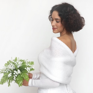 Puede incluir: Un chal de novia de punto blanco con un escote drapeado. El chal se lleva sobre un vestido blanco y la modelo sostiene un ramo de flores blancas.