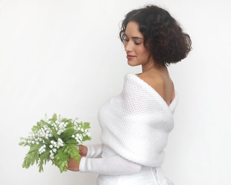 Puede incluir: Un chal de novia de punto blanco con un escote drapeado. El chal se lleva sobre un vestido blanco y la modelo sostiene un ramo de flores blancas.