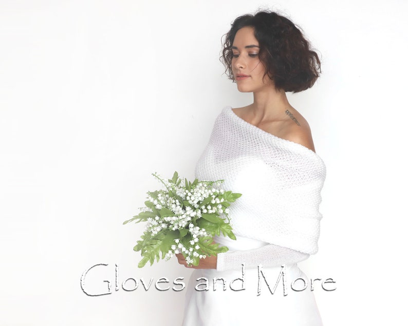 Puede incluir: Un su&eacute;ter blanco de punto con hombros descubiertos y mangas largas. El su&eacute;ter est&aacute; drapeado sobre los hombros y se usa con un vestido blanco. La modelo sostiene un ramo de flores blancas. El texto "Gloves and More" est&aacute; escrito en negro en la parte inferior de la imagen.