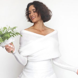 Puede incluir: Una estola blanca de punto de hombros descubiertos con mangas largas. La estola est&aacute; drapeada sobre los hombros y tiene un corte holgado y fluido. La modelo sostiene un ramo de flores blancas.