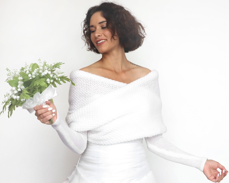Puede incluir: Una estola blanca de punto de hombros descubiertos con mangas largas. La estola est&aacute; drapeada sobre los hombros y tiene un corte holgado y fluido. La modelo sostiene un ramo de flores blancas.