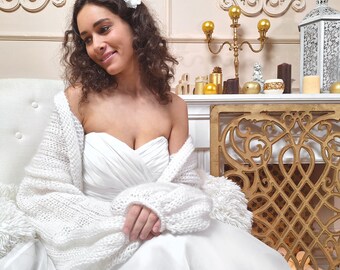 May include: A white bridal gown with a sweetheart neckline and a matching white knitted shawl. The shawl has long sleeves and is draped over the shoulders. A white flower hair accessory is visible. The background includes a fireplace and decorative items.