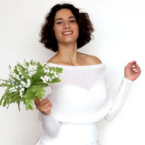 Puede incluir: Una estola de novia blanca de punto con hombros descubiertos y mangas largas. La modelo sostiene un ramo de flores blancas de lirio de los valles.