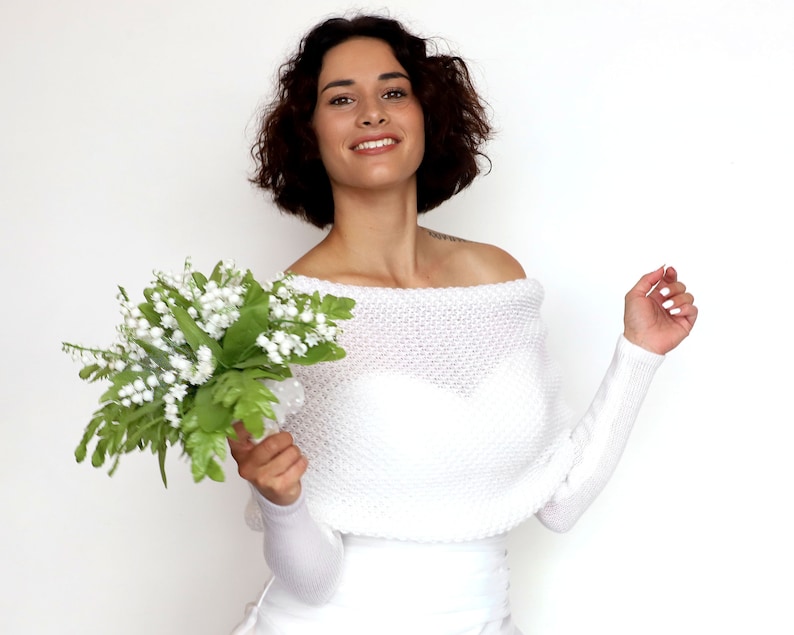 Puede incluir: Una estola de novia blanca de punto con hombros descubiertos y mangas largas. La modelo sostiene un ramo de flores blancas de lirio de los valles.