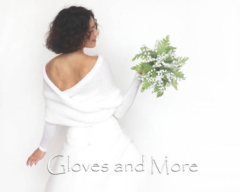 Puede incluir: Una estola blanca de punto con mangas largas y escote drapeado. La estola se lleva sobre un vestido blanco. La modelo sostiene un ramo de flores blancas con hojas verdes. El texto "Gloves and More" es visible en la parte inferior de la imagen.