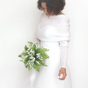 Puede incluir: Una estola de novia de punto blanco con mangas largas y escote drapeado. La estola se lleva sobre un vestido de novia blanco y la modelo sostiene un ramo de flores blancas.
