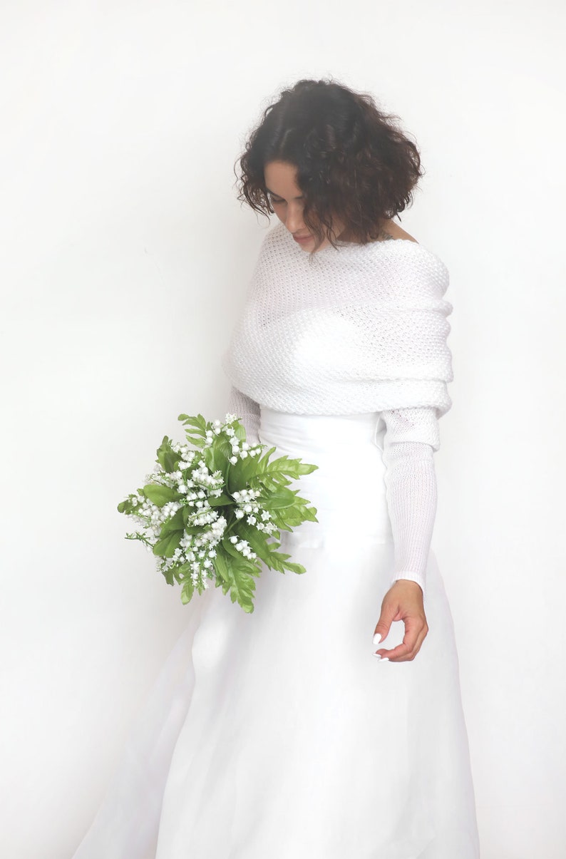 Puede incluir: Una estola de novia de punto blanco con mangas largas y escote drapeado. La estola se lleva sobre un vestido de novia blanco y la modelo sostiene un ramo de flores blancas.