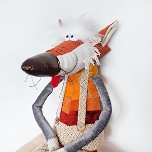 May include: A quirky stuffed wolf toy with a long snout, white fur, and a red tongue. The wolf is dressed in a patchwork vest in shades of orange and red, with grey arms and legs. The toy has button details and a playful expression.
