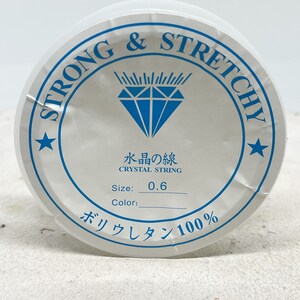 Puede incluir: Una bobina blanca de hilo de cristal fuerte y el&aacute;stico, etiquetada como "Strong & Stretchy" y "Crystal String". La bobina est&aacute; etiquetada con el tama&ntilde;o "0,6" y el material "Poliuretano 100&nbsp;%".